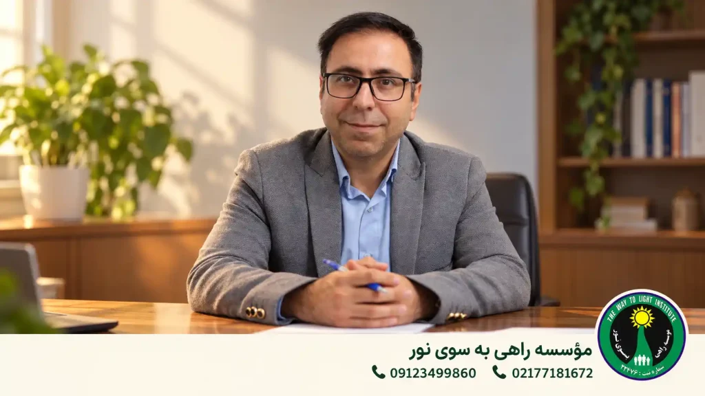 مردی با کت خاکستری و عینک پشت میز چوبی در دفتر با نور ملایم و گیاهان سبز