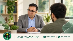 روان‌شناس با کت خاکستری در حال گفت‌وگو با یک مراجعه‌کننده در اتاق روشن مشاوره
